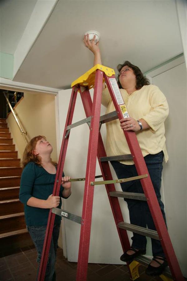 Woman on ladder installing smoke detector while daughter stabilizes ladder